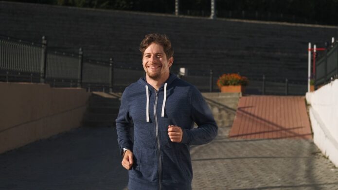 A smiling man in a navy blue hoodie jogs outside on a paved path with steps and flower pots in the background on a sunny day