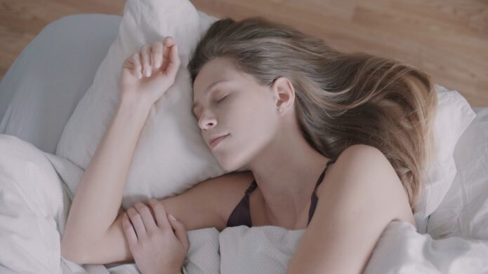 A young woman with long blonde hair sleeps on a white bed, resting on her side with a peaceful expression