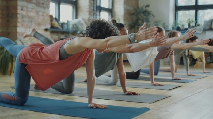 A diverse group of people practicing yoga in a well-lit studio, balancing on one leg and extending their arms in a stretching pose