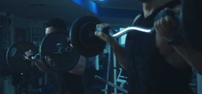 Two men in black sleeveless shirts performing barbell curls in a dimly lit gym with blue lighting