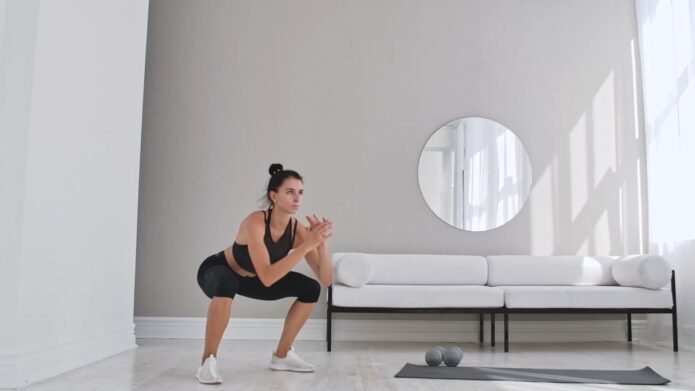 A woman in a black sports outfit performs a squat exercise in a bright, minimalist living room with a white couch, round mirror, and yoga mat