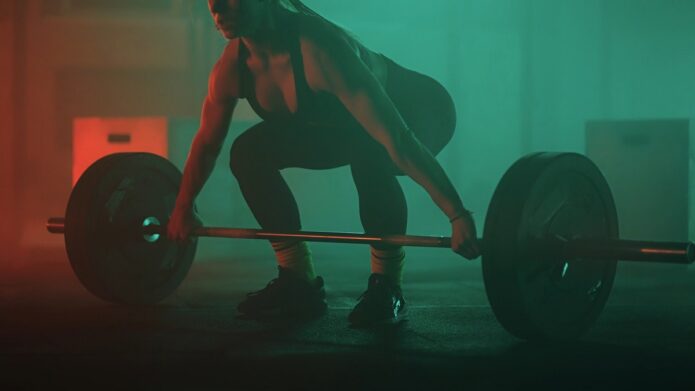 A fit woman in a black workout outfit crouches to lift a barbell in a dimly lit gym with green and red lighting