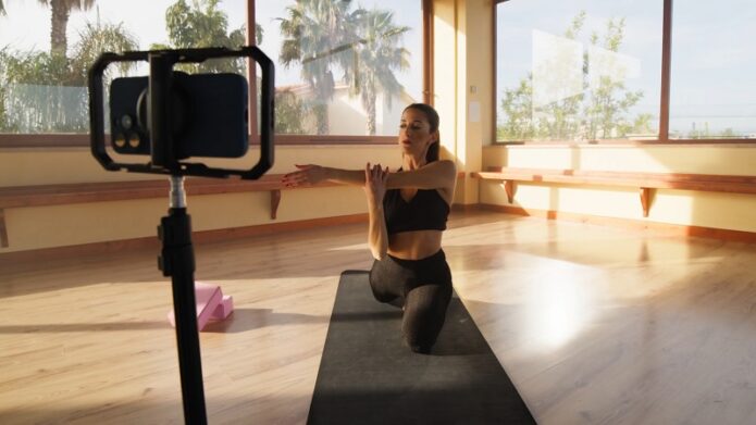 A fitness instructor records workout clips on her smartphone in a sunlit yoga studio
